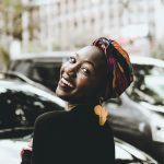 A joyful woman wearing a colorful headwrap and distinctive earrings, smiling in an urban setting.