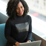 Beautiful African American woman with curly hair smiling while using a laptop indoors.