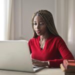 Confident woman in a red sweater working on a laptop in a bright, organized workspace.