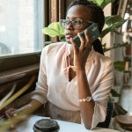 Young woman with eyeglasses talking on smartphone at cafe table with coffee, notebook, and plants.